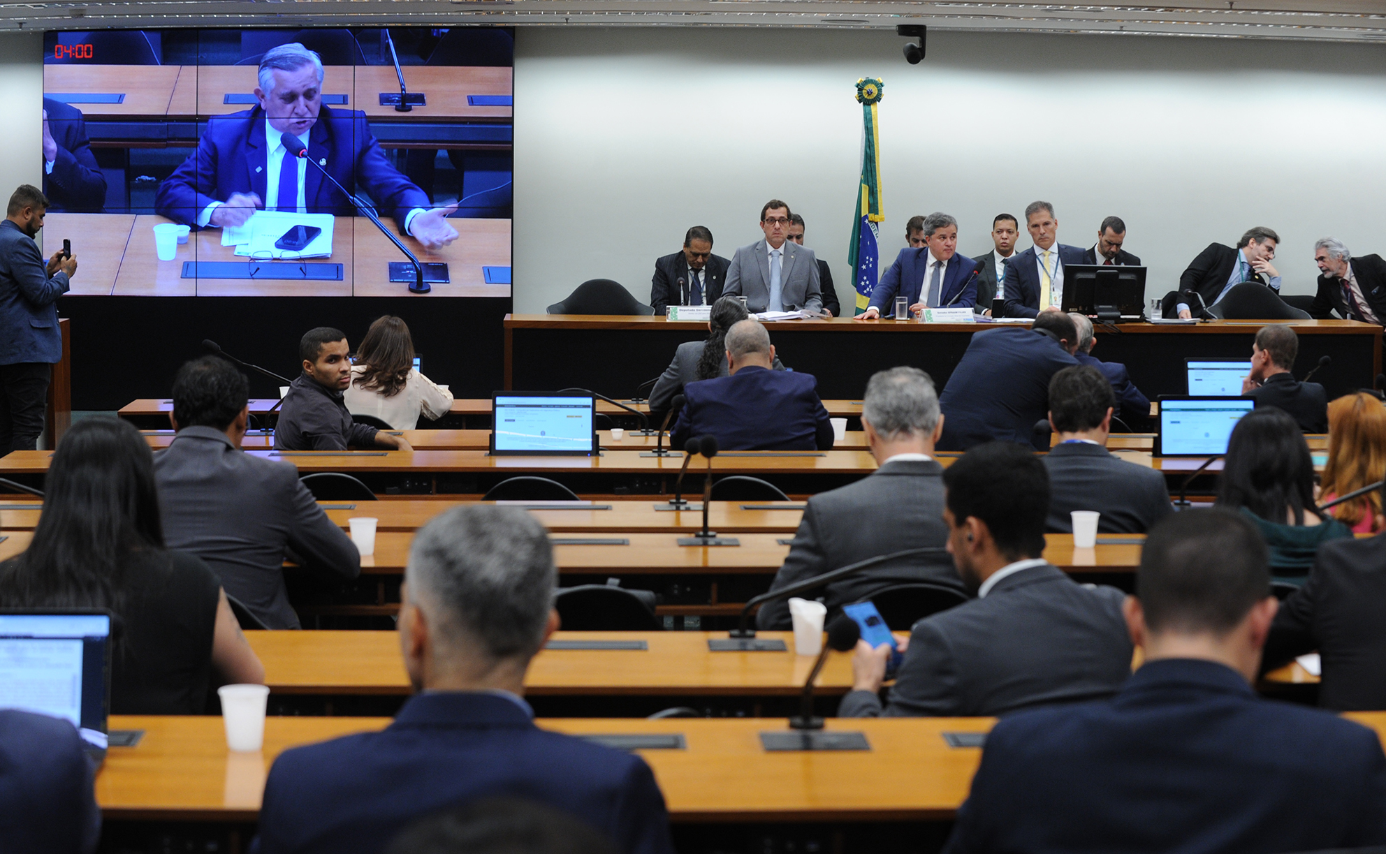 Foto de sessão da Comissão Mista de Orçamento. Mesas enfileiradas com laptops sobre elas, e alguns parlamentares ocupando alguns assentos. À frente, a mesa diretora.
